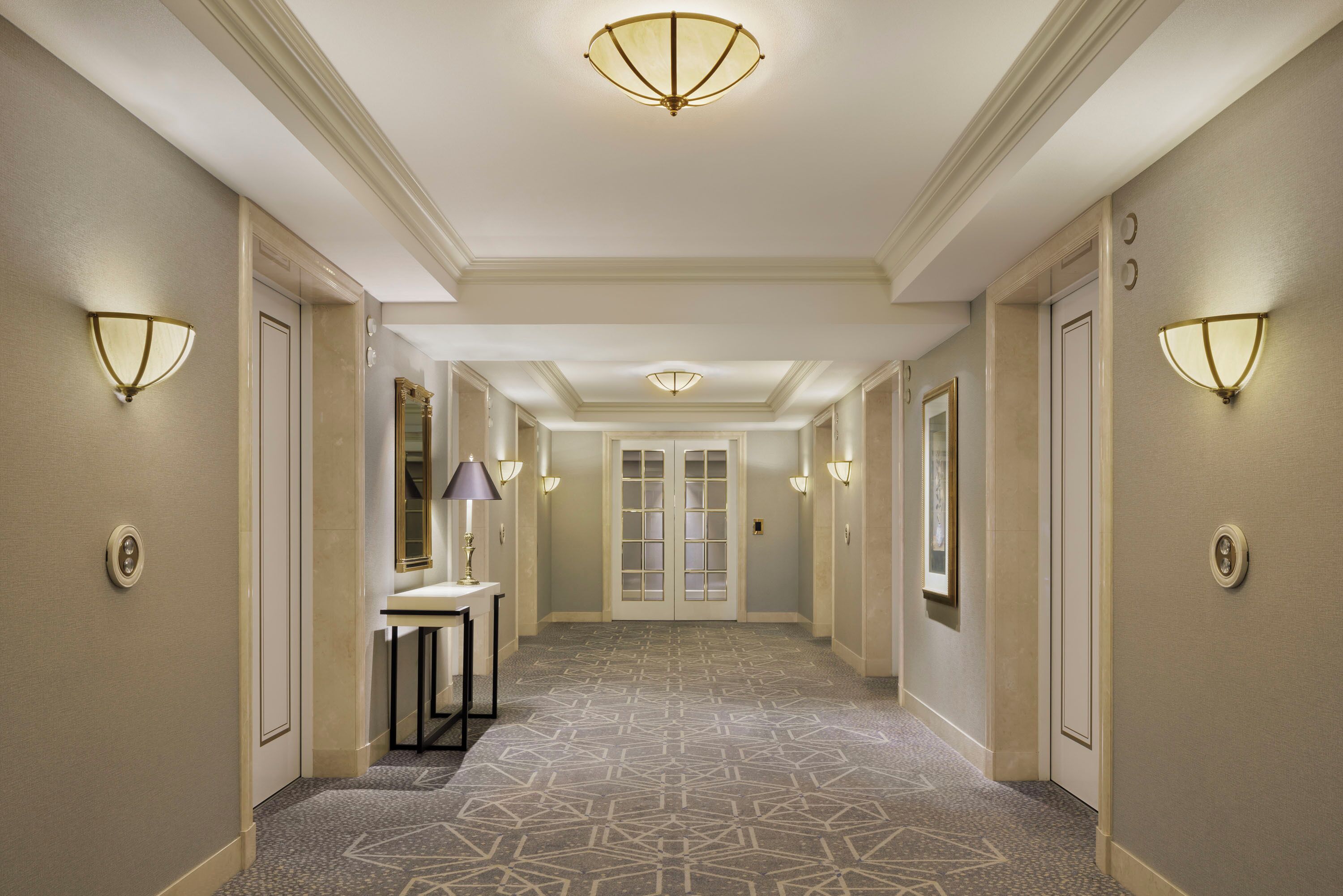 A hotel hallway with patterned carpet, wall sconces, and a central door with glass panels.
