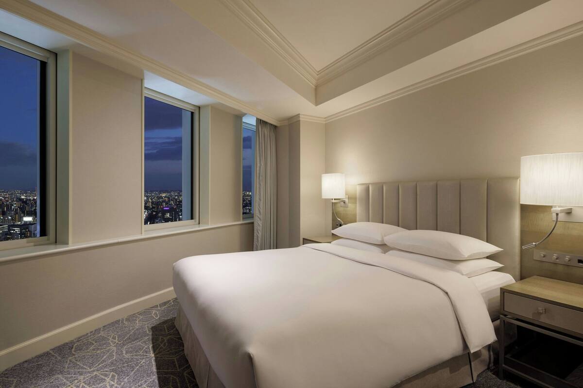 A hotel room with a large bed, a nightstand, a lamp, and a view of a cityscape at night.