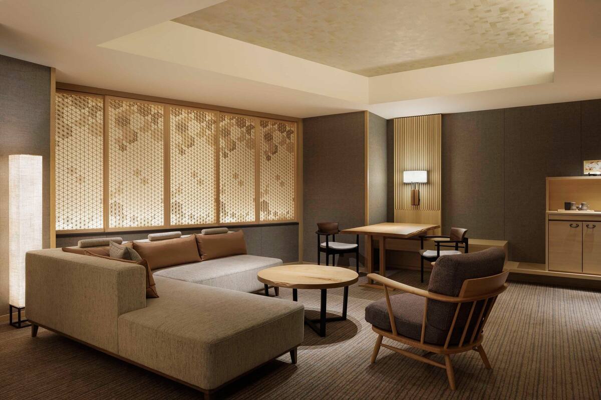 A modern hotel room with a sectional sofa, a round coffee table, a wooden chair, a small dining table, and a kitchen area.