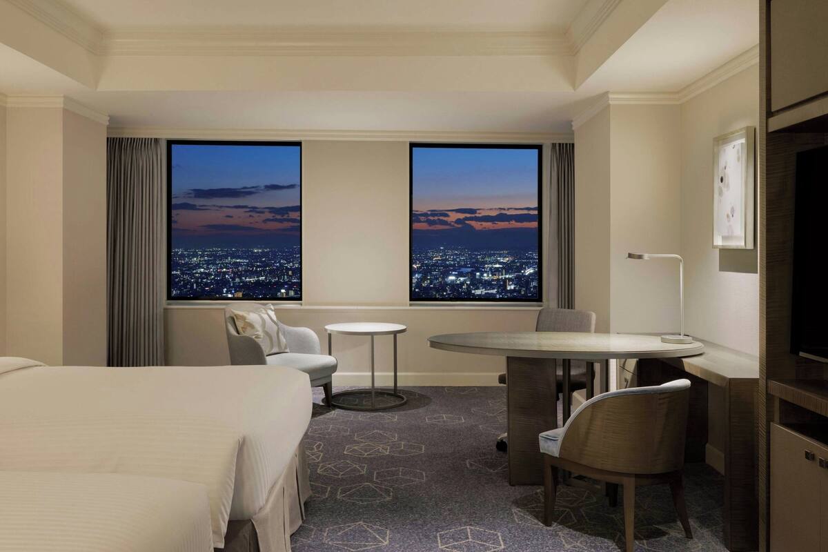 A hotel room with a large bed, a desk, two chairs, a small table, and a view of a cityscape at dusk.