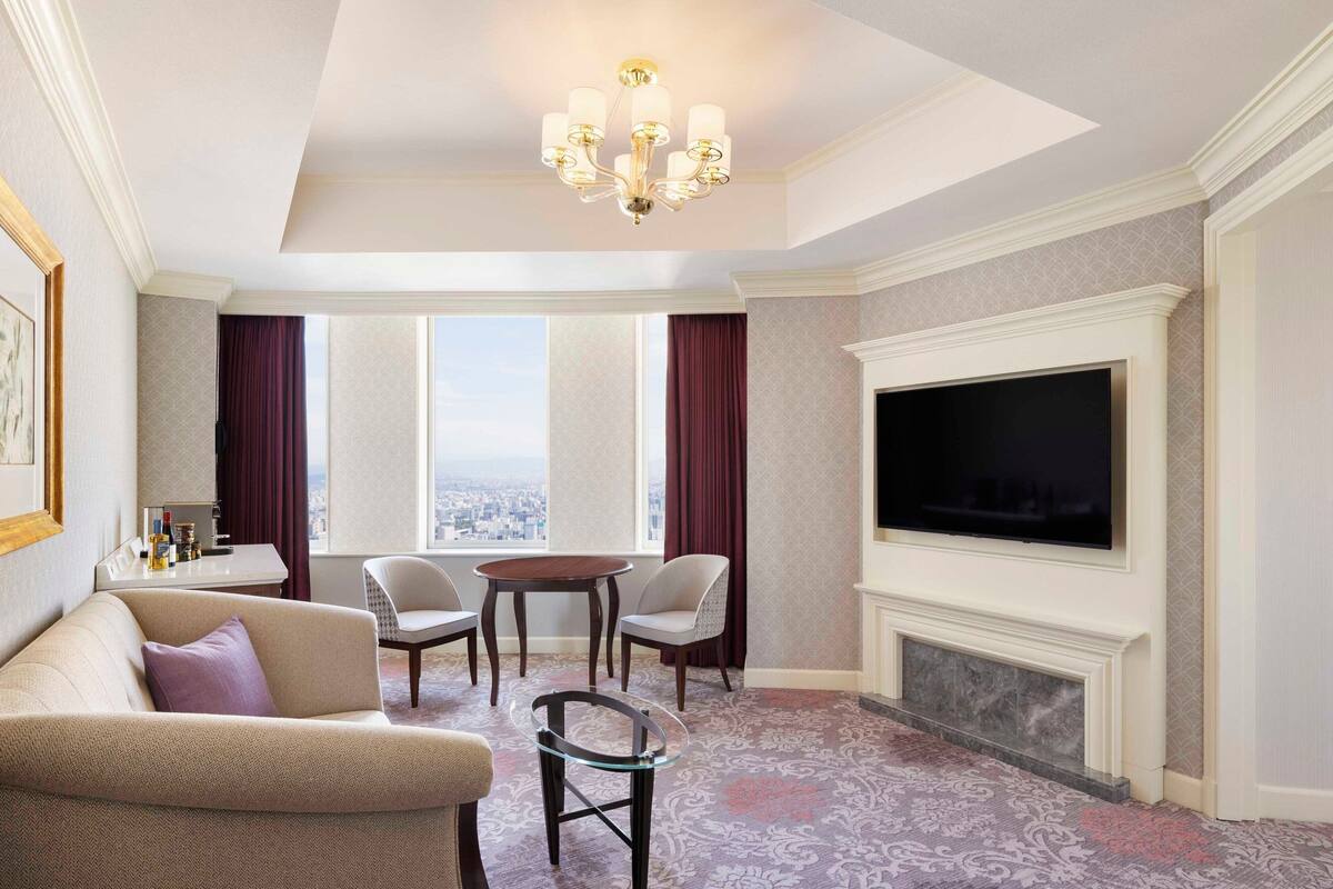 A hotel room with a large flat-screen TV, a fireplace, a sofa, a dining table with chairs, and a view of the city through large windows.