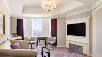 A hotel room with a large flat-screen TV, a fireplace, a sofa, a dining table with chairs, and a view of the city through large windows.