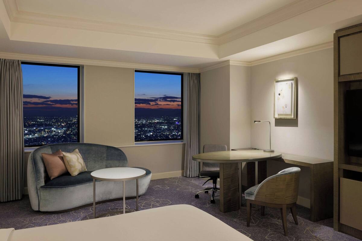 A hotel room with a city view, a sofa, a small table, a desk, and a chair.