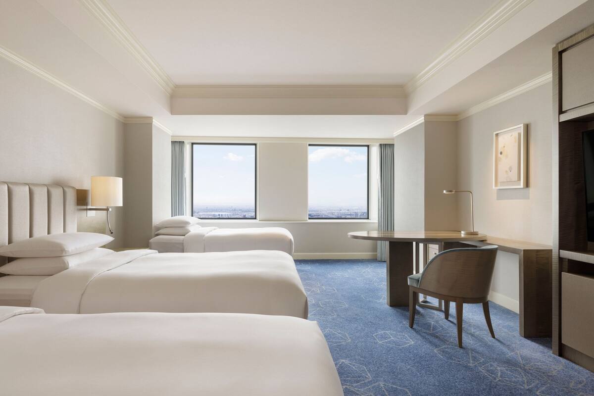 A hotel room with two beds, a desk, a chair, and a large window offering a city view.