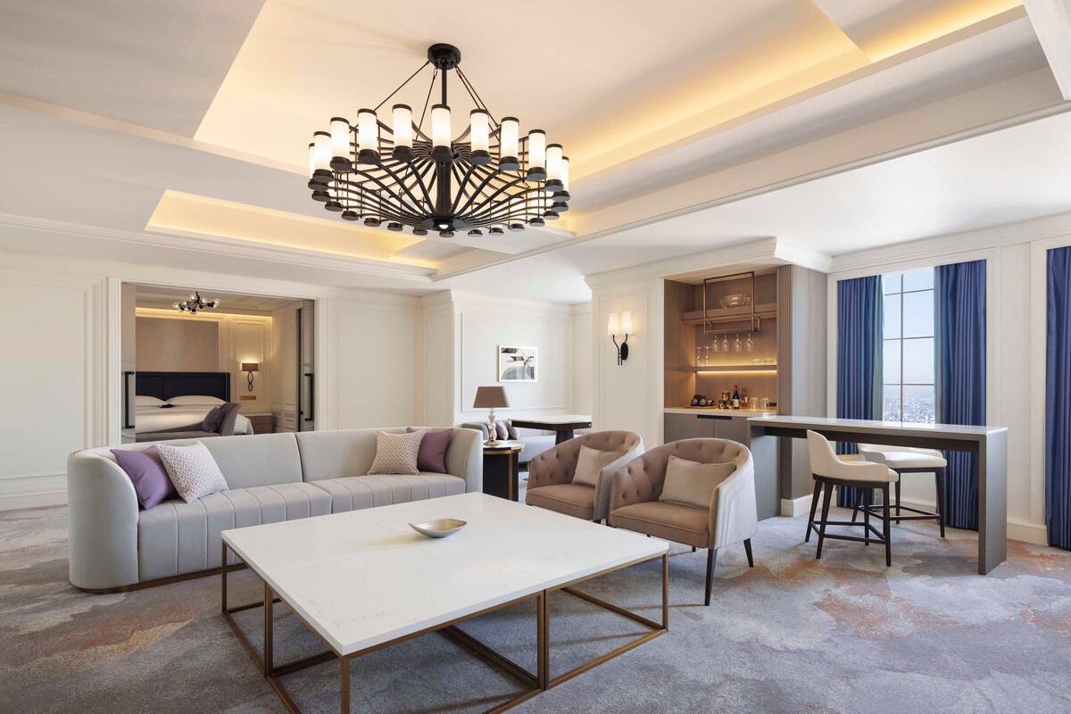 A spacious living room with a modern chandelier, a sofa set, a dining area with bar stools, and a view of the city through large windows.