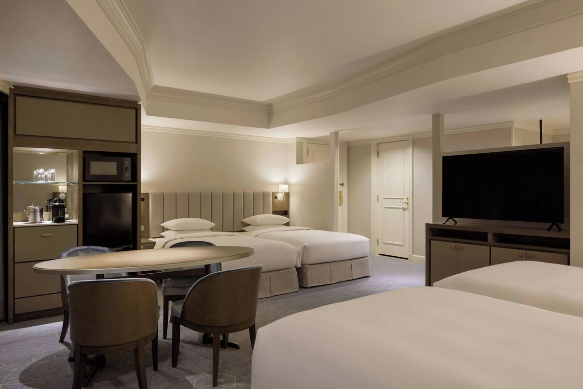 A hotel room with two beds, a desk with chairs, a television, and a minibar.