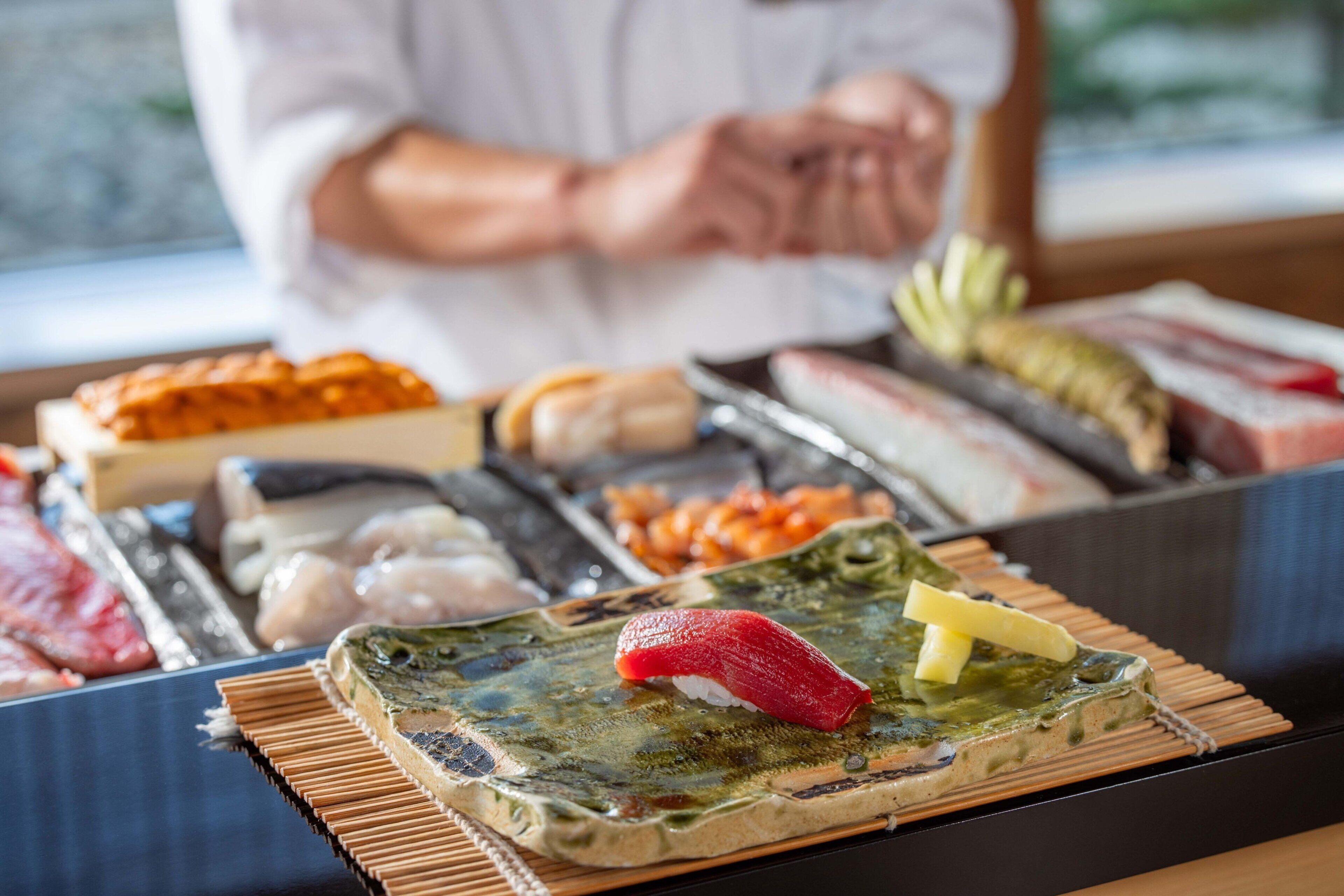 A sushi platter with various types of fish and toppings, a chef in the background, and a wooden table.