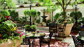 An outdoor patio area with tables and chairs, surrounded by potted plants and a fountain.