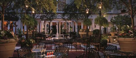 An outdoor patio area with tables and chairs, surrounded by potted plants and trees.