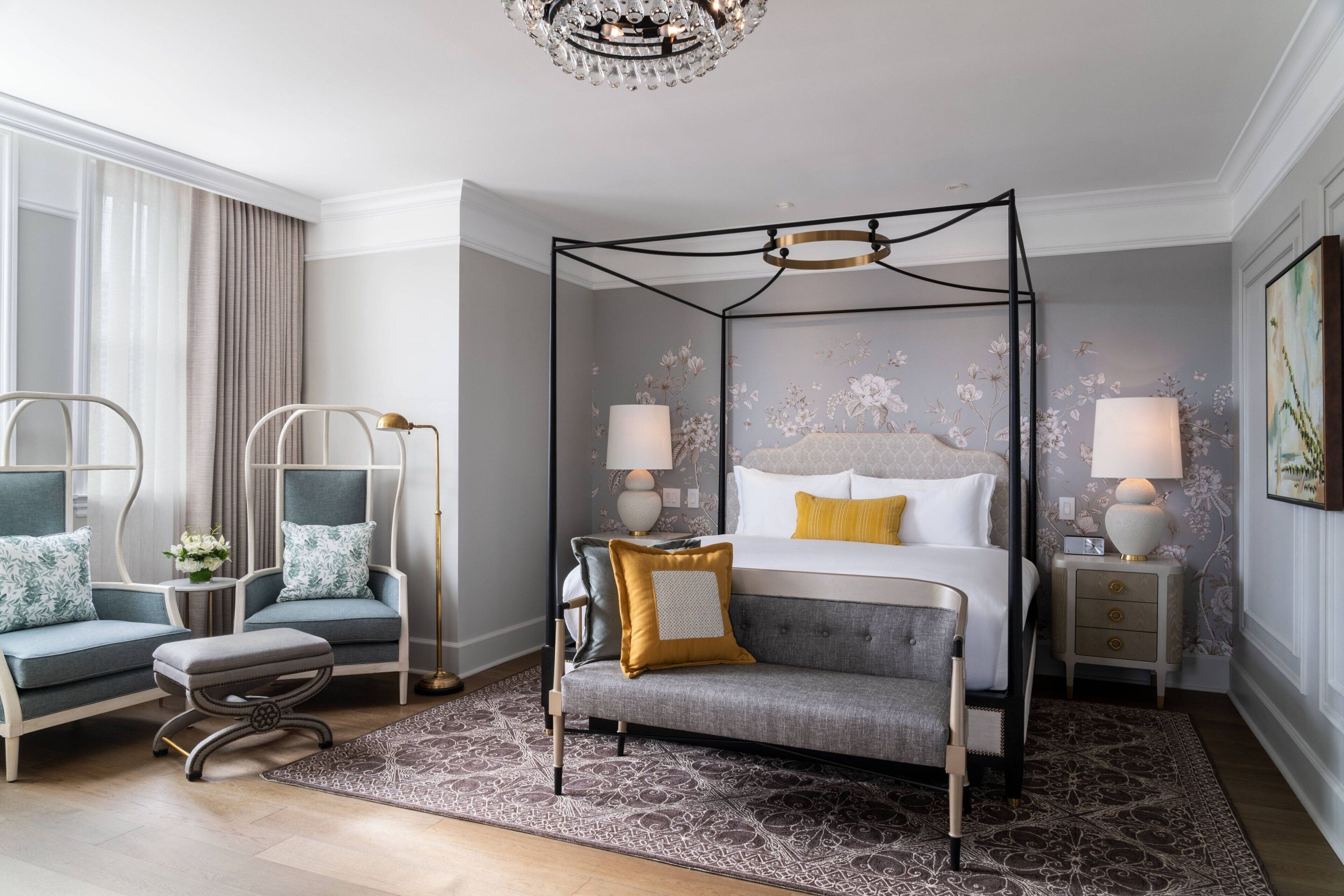 A modern bedroom with a four-poster bed, two armchairs, a bench, and a nightstand with lamps.
