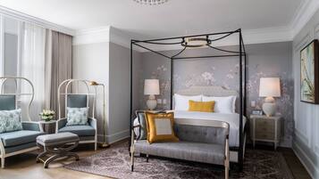 A modern bedroom with a four-poster bed, two armchairs, a bench, and a nightstand with lamps.