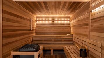 Una sauna con bancos de madera y un asiento, un cubo negro, y un banco con cojín negro.