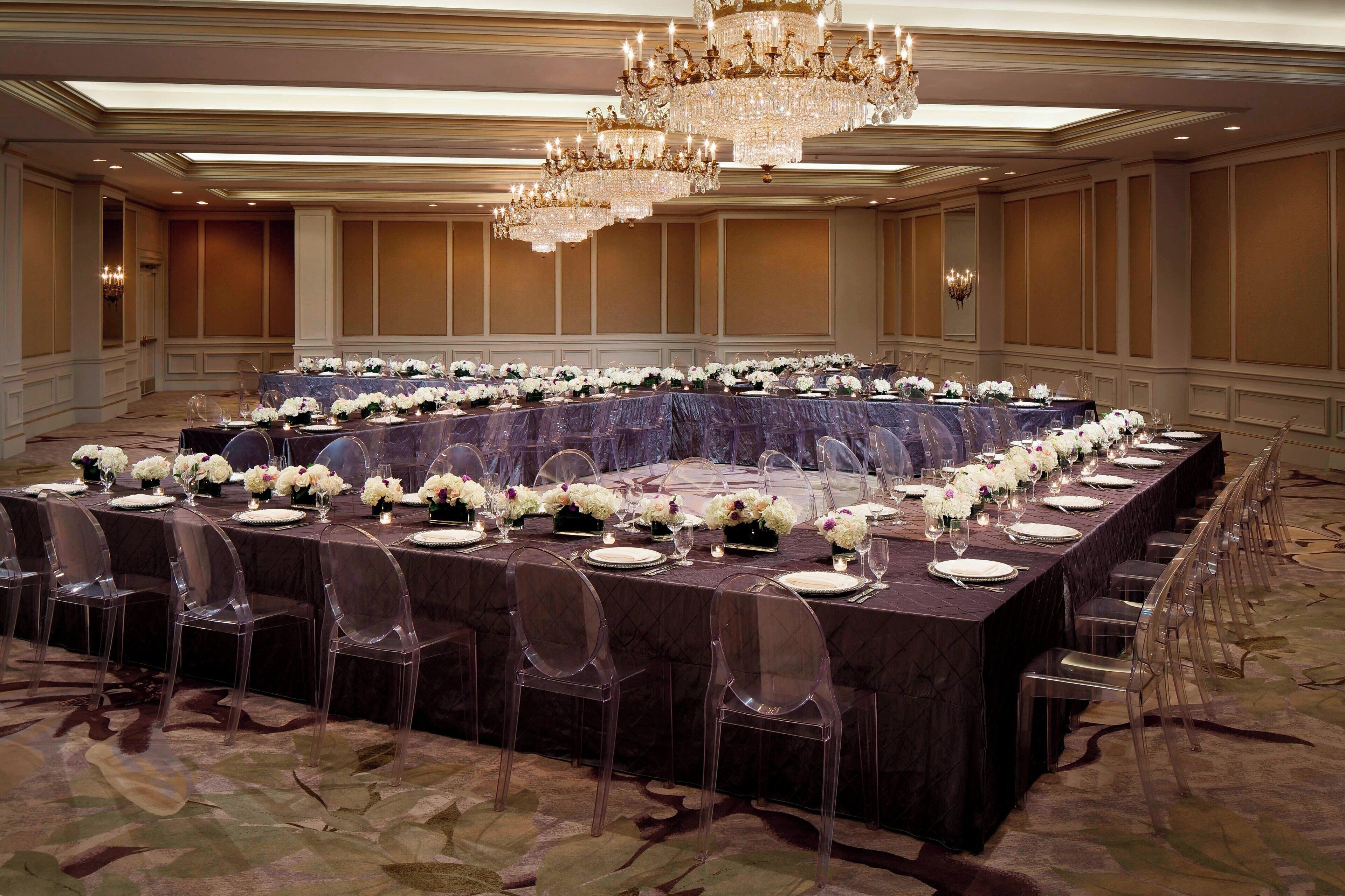 A banquet hall with a long rectangular table set for a formal event, surrounded by clear chairs, and adorned with white floral arrangements.