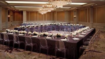 A banquet hall with a long rectangular table set for a formal event, surrounded by clear chairs, and adorned with white floral arrangements.