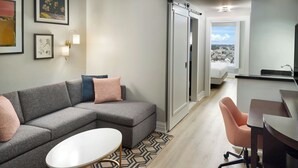 Una habitación de hotel moderna con un sofá seccional gris, una pequeña mesa redonda, un escritorio con una silla, una cama, un baño con lavamanos y vistas a la ciudad a través de una ventana.