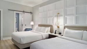 Habitación de hotel con dos camas, una mesita de noche con lámpara, una silla y una puerta que da al baño.