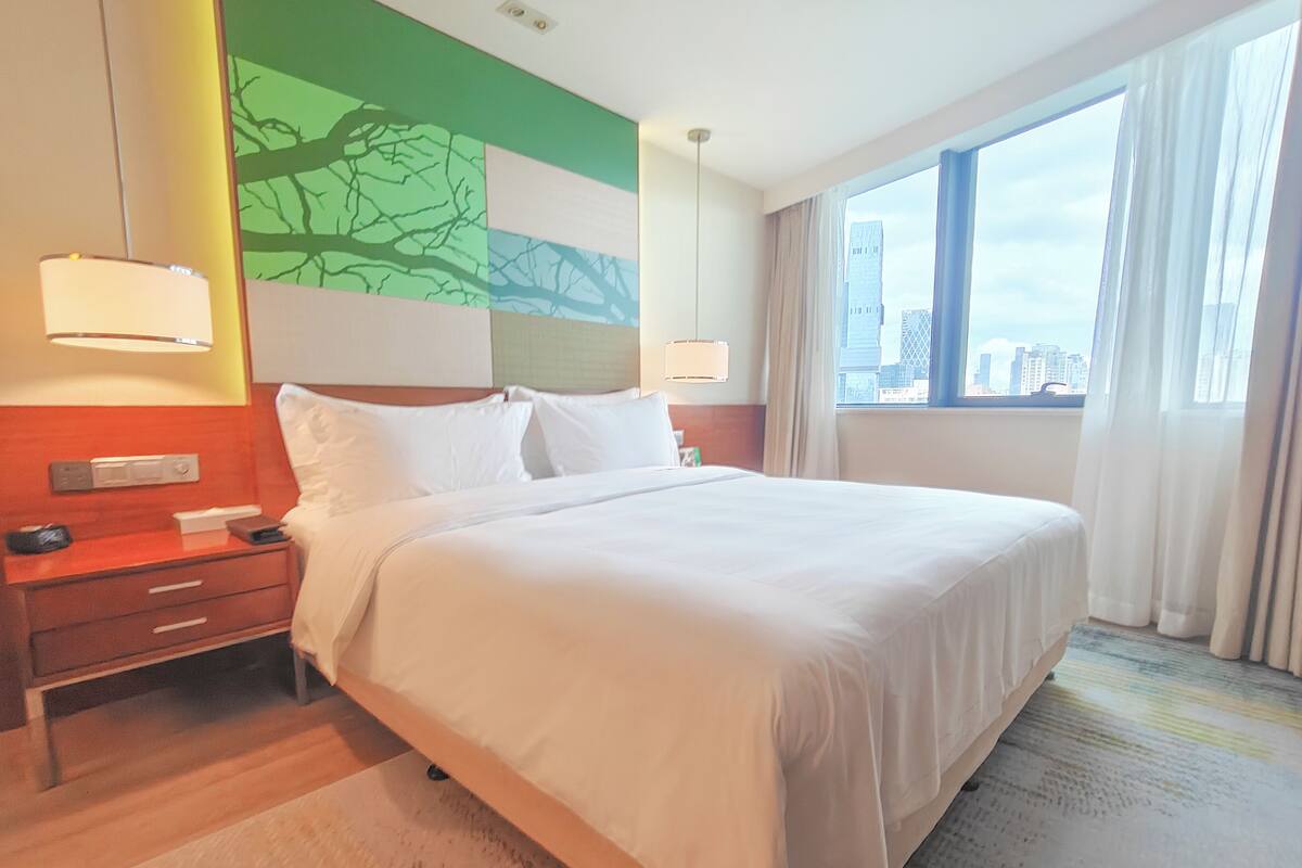 A hotel room with a large bed, a nightstand, a wall-mounted lamp, and a window with a city view.
