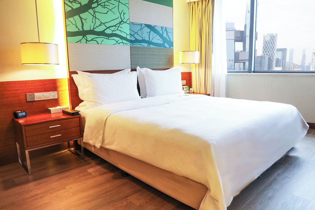 A hotel room with a large bed, two bedside tables, a nightstand, a wall-mounted lamp, and a view of the city skyline through the window.