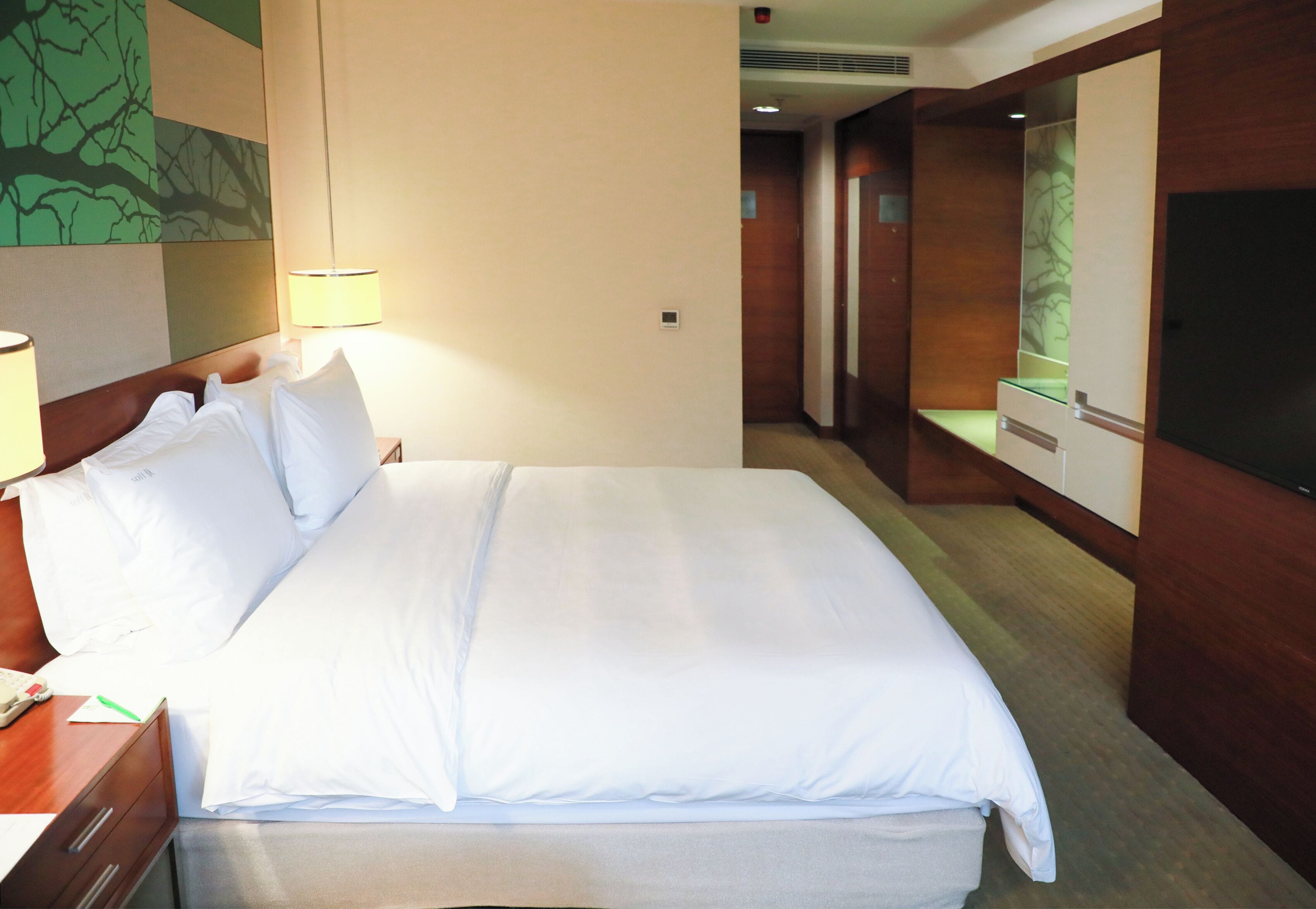 A hotel room with a neatly made bed, a bedside lamp, a wooden nightstand, and a large flat-screen TV mounted on the wall.