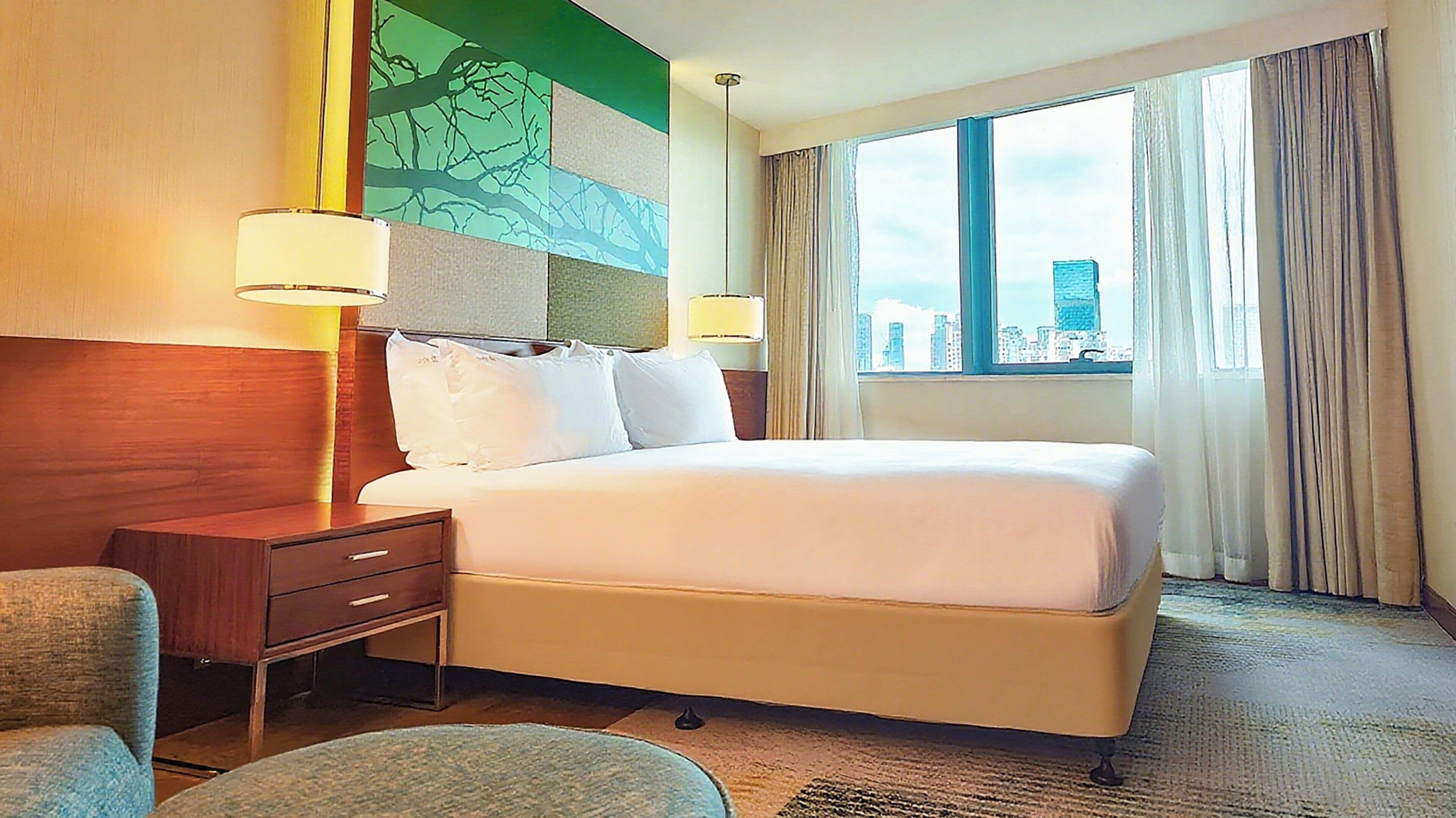 Quarto de hotel com uma cama grande, duas mesas de cabeceira com luminárias, uma cadeira e vista para a cidade pela janela.