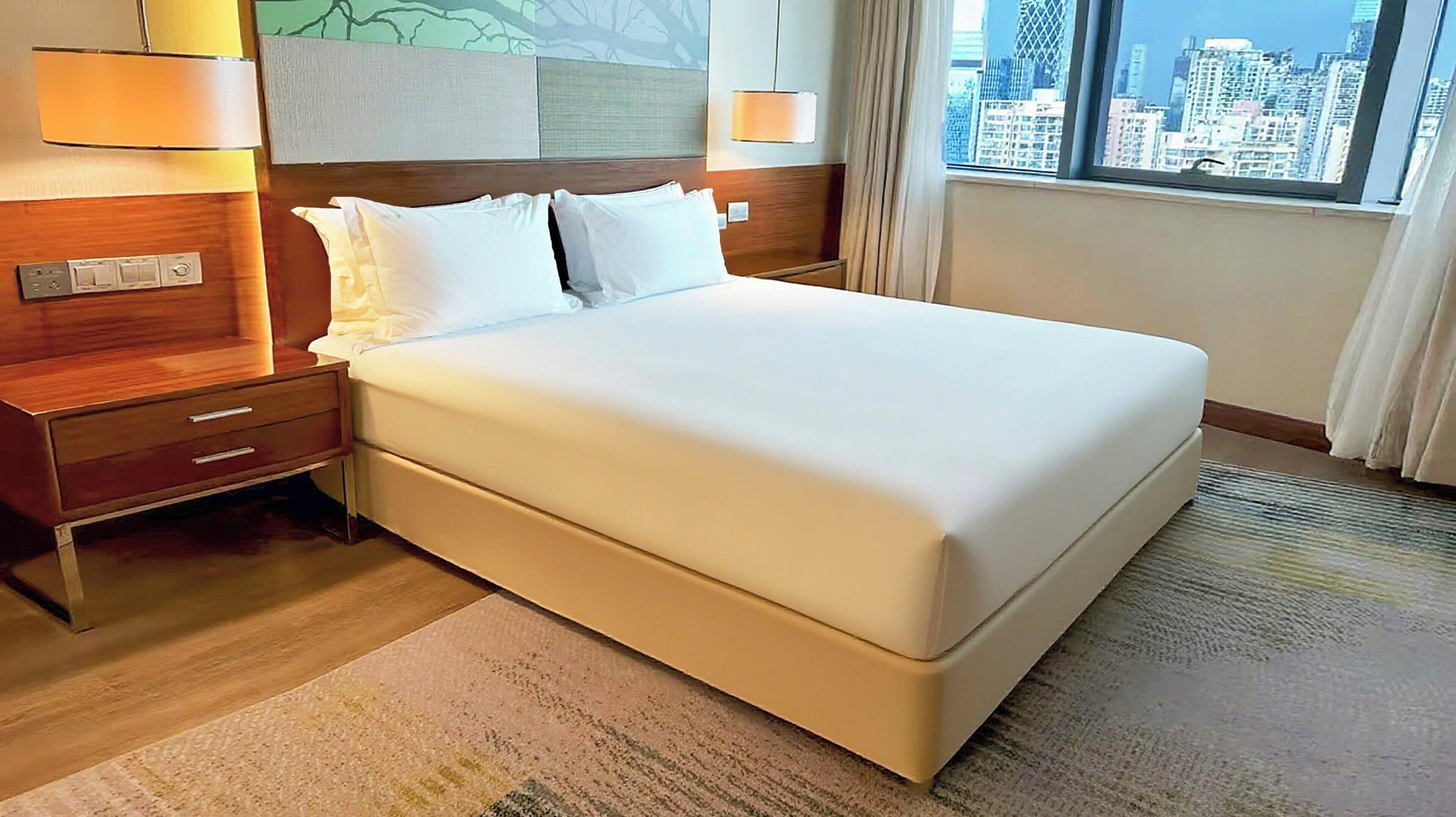 A hotel room with a large bed, wooden headboard, bedside lamps, and a view of the city through the window.