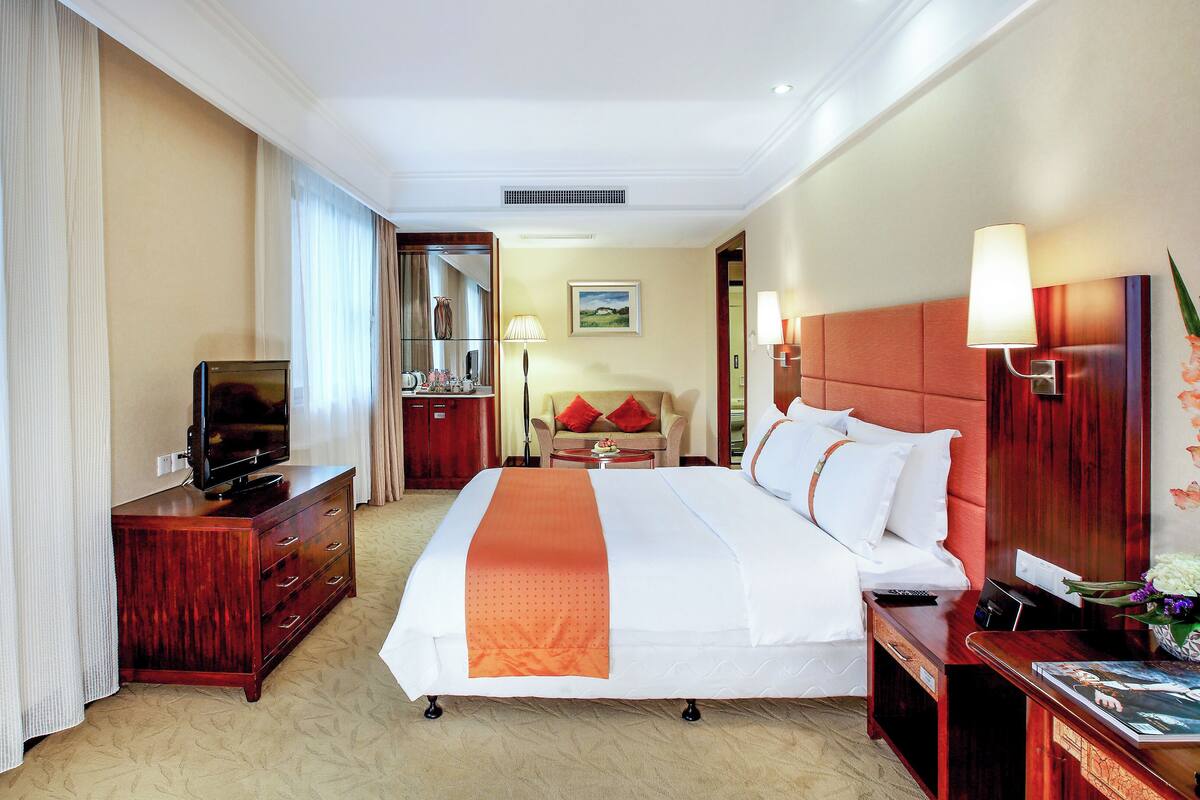 A hotel room with a bed, a TV on a wooden dresser, a desk with a lamp, and a view of the hallway.