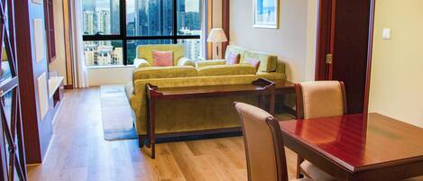 A hotel room with a large window offering a city view, a green sofa, a wooden dining table, and a chandelier.