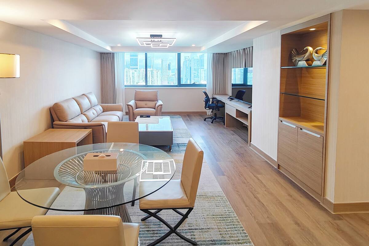 A modern hotel room with a glass dining table, leather sofas, a wooden desk, and a large window offering a city view.