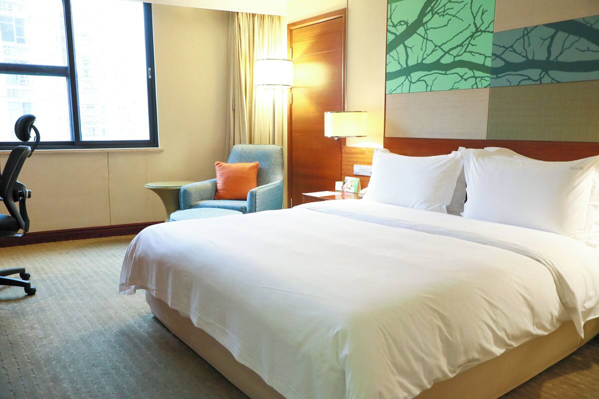 A hotel room with a large bed, a desk with a chair, a lamp, and a window with curtains.