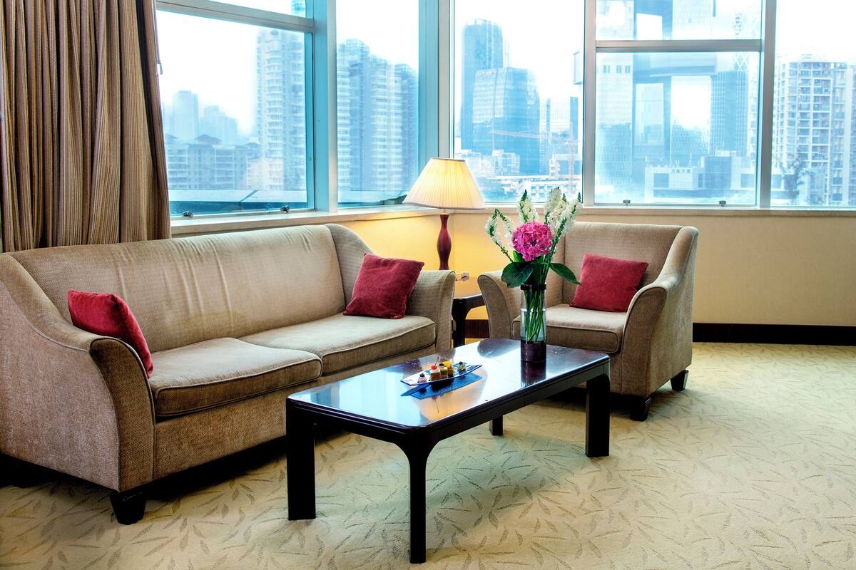 A modern hotel room with a sofa set, a coffee table with a vase of flowers, and a view of the cityscape through large windows.