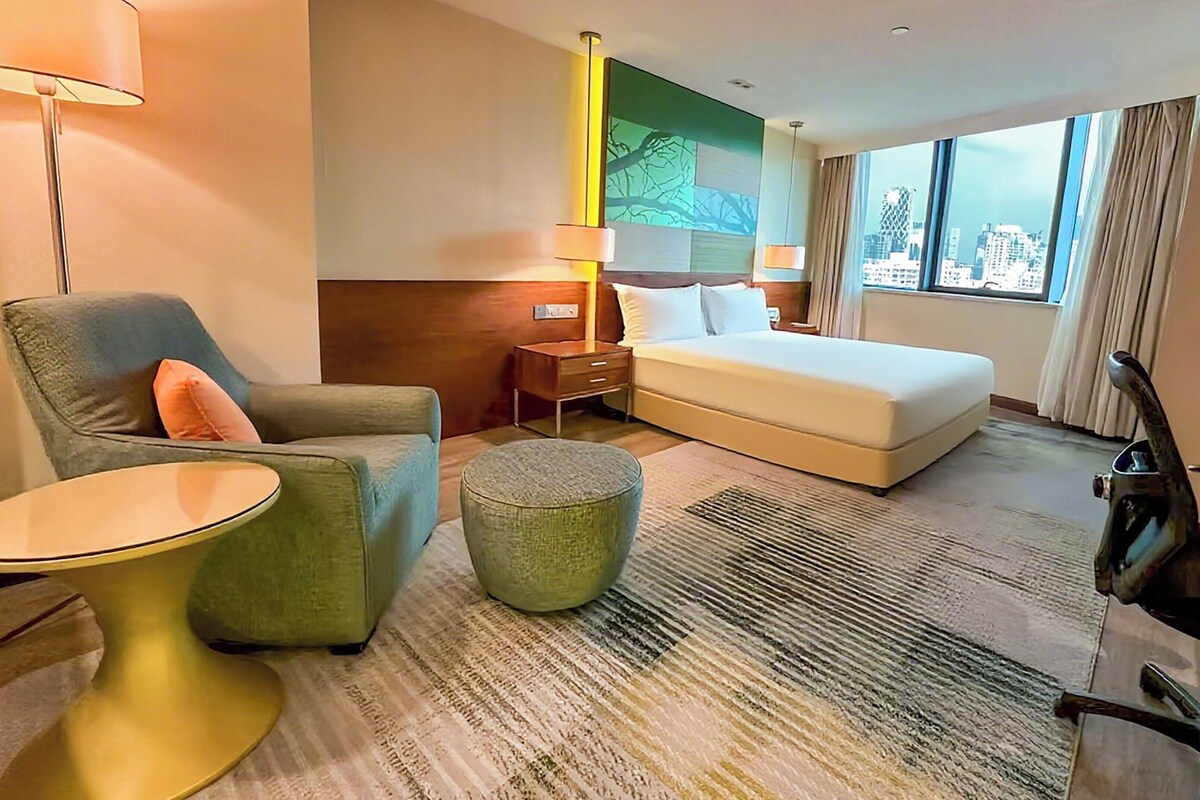 A hotel room with a bed, a chair, a small table, a lamp, and a view of the city through large windows.