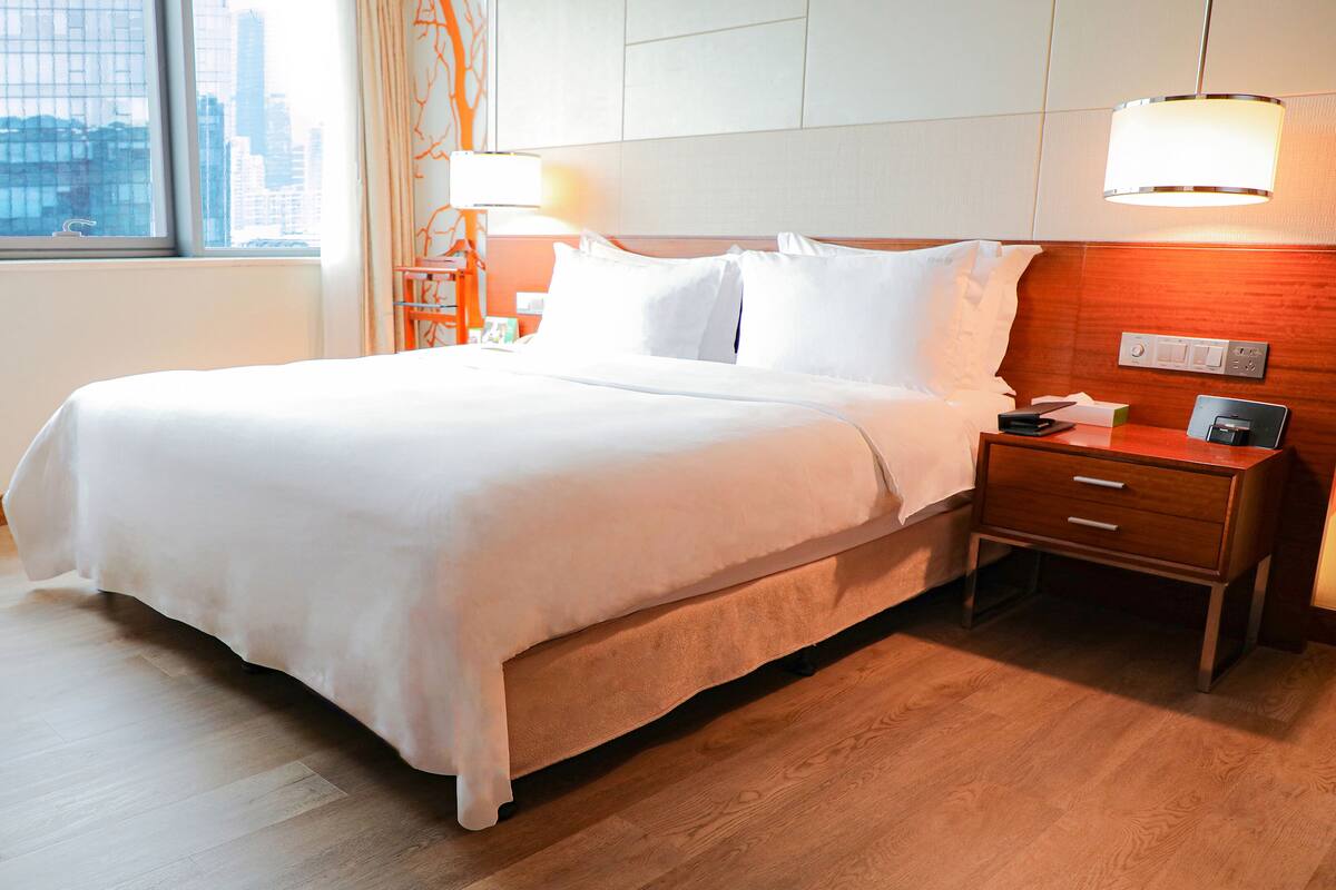 A neatly made bed with white linens, a wooden nightstand with a lamp, and a view of the city through a window.