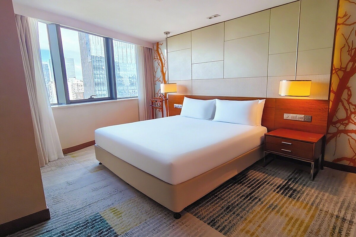 A hotel room with a large bed, a nightstand with a lamp, a window with curtains, and a view of the cityscape.