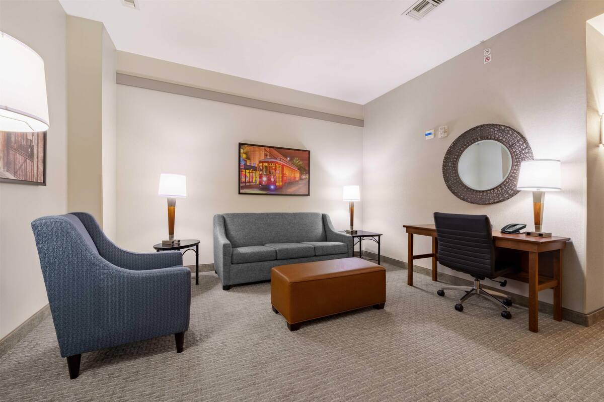 A hotel room with a gray sofa, a blue armchair, a wooden coffee table, a desk with a chair, a round mirror, and a painting on the wall.
