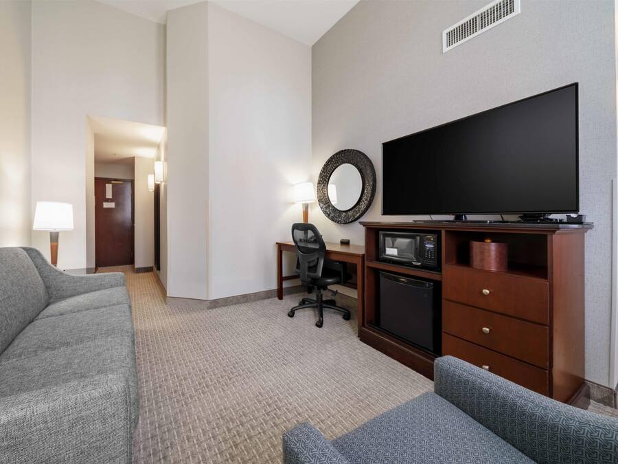 A hotel room with a gray sofa, a smaller gray armchair, a desk with a chair, a flat-screen TV, a microwave, and a wooden cabinet.