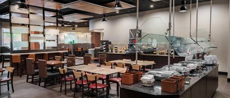 Un restaurante con varias mesas y sillas, una barra de buffet con recipientes de comida y una zona de cocina al fondo.