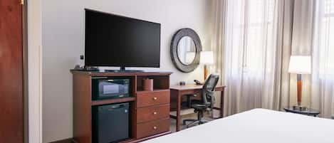 A hotel room with a bed, a flat-screen TV, a microwave, a refrigerator, a desk with a chair, and a large mirror.