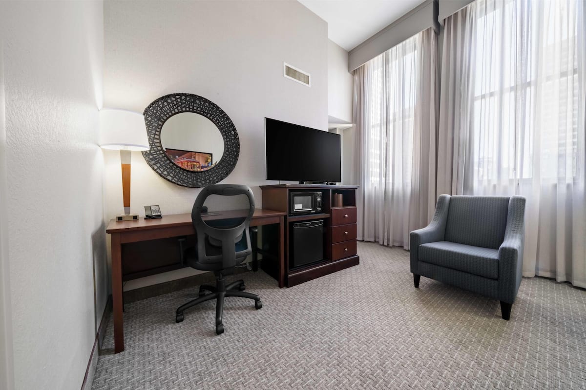 A hotel room with a desk, chair, television, and a large window with curtains.