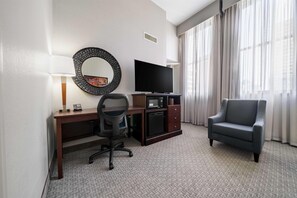 Habitación de hotel con escritorio, silla, televisión y un amplio ventanal con cortinas.