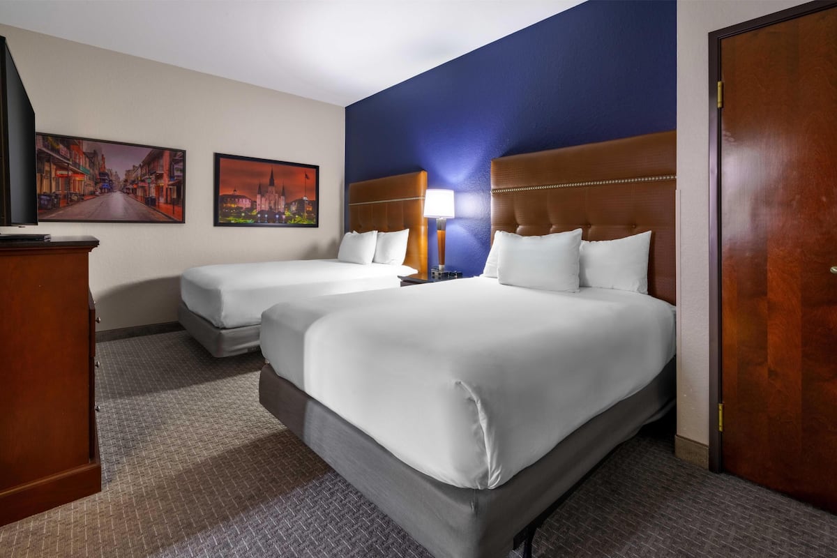 A hotel room with two beds, a wooden headboard, a desk, a lamp, and two framed pictures on the wall.