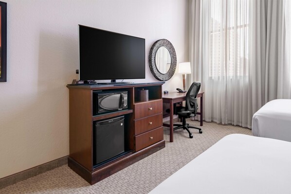 Habitación de hotel con televisión de pantalla plana, escritorio con silla, refrigerador pequeño, espejo y cama.