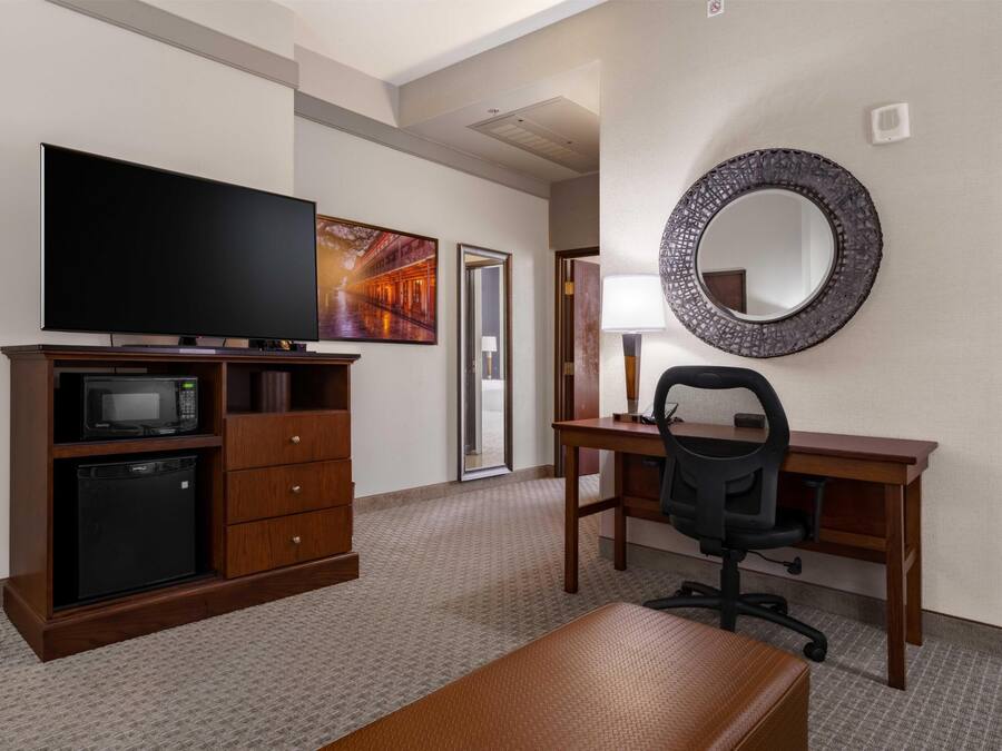 Habitación de hotel con televisor de pantalla plana, microondas, refrigerador pequeño, escritorio de madera, silla de oficina negra, espejo redondo y una pintura grande en la pared.
