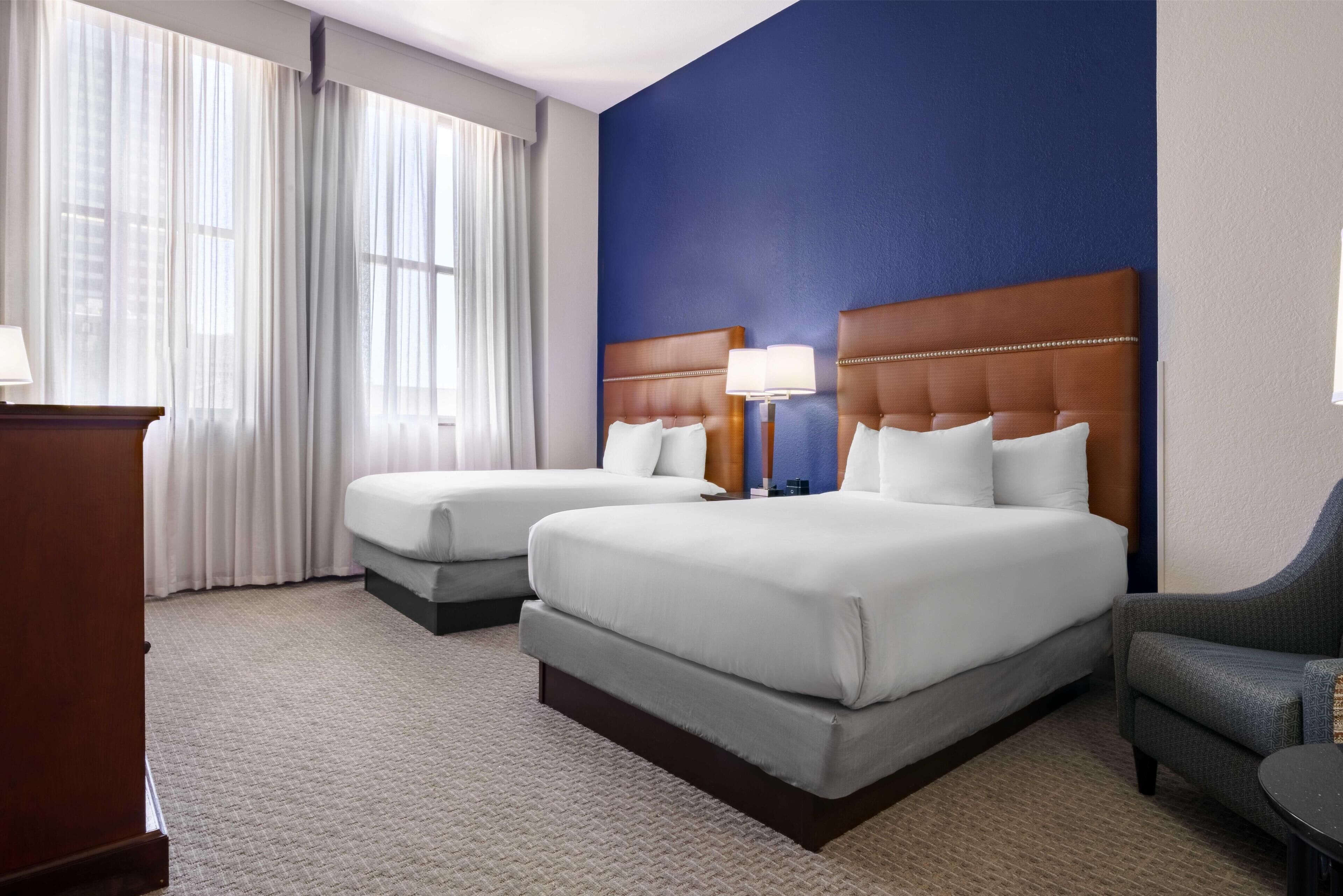 A hotel room with two beds, a desk, a chair, and a large window with curtains.