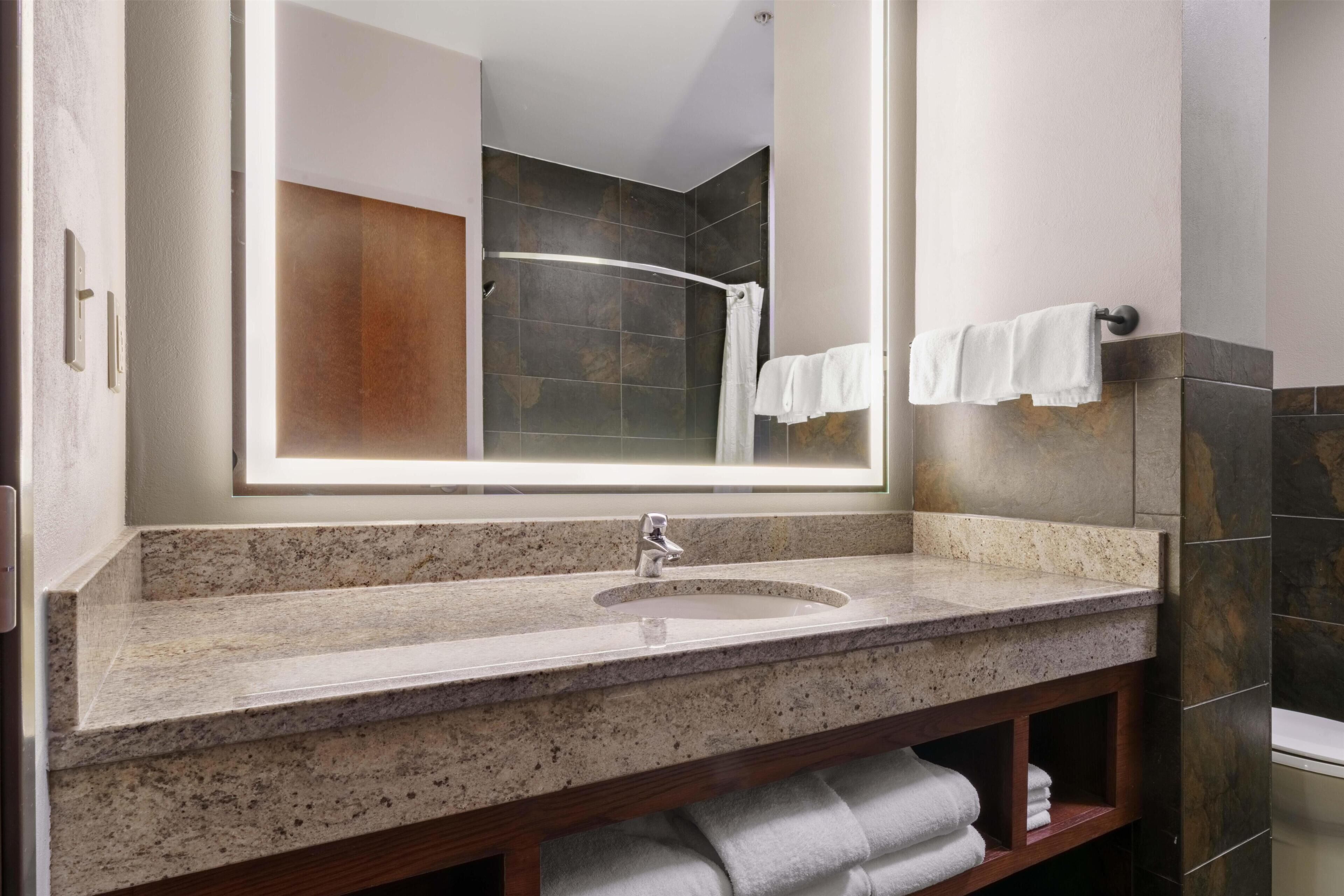 A bathroom with a granite countertop sink, a large mirror, a shower, and a towel rack.