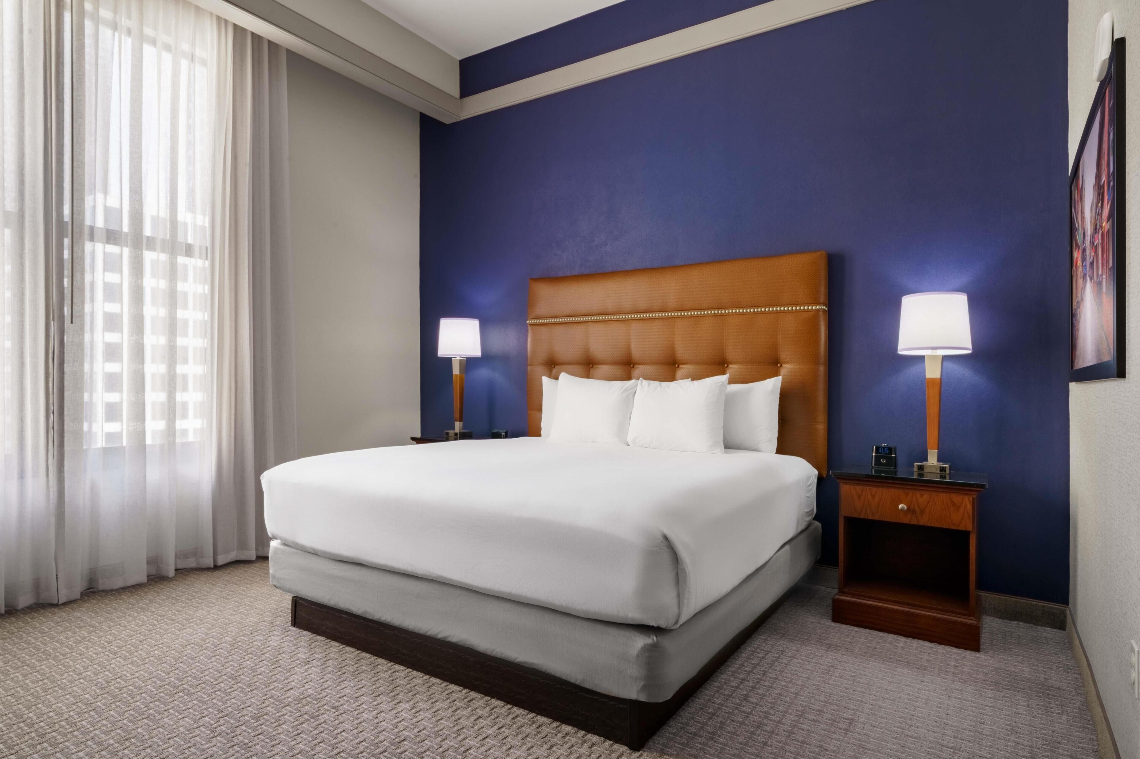 Una habitación de hotel con una cama grande, dos mesitas de noche con lámparas, una ventana con cortinas translúcidas y una pared de acento azul oscuro.