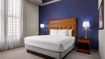 Una habitación de hotel con una cama grande, dos mesitas de noche con lámparas, una ventana con cortinas translúcidas y una pared de acento azul oscuro.