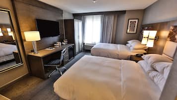 Habitación de hotel con dos camas, un escritorio con lámpara, televisión y un ventanal grande con cortinas.