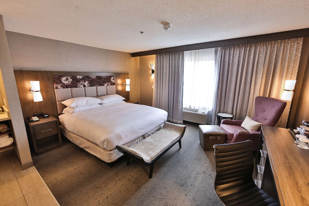 A hotel room with a large bed, bedside tables, a desk, a chair, and a window with curtains.