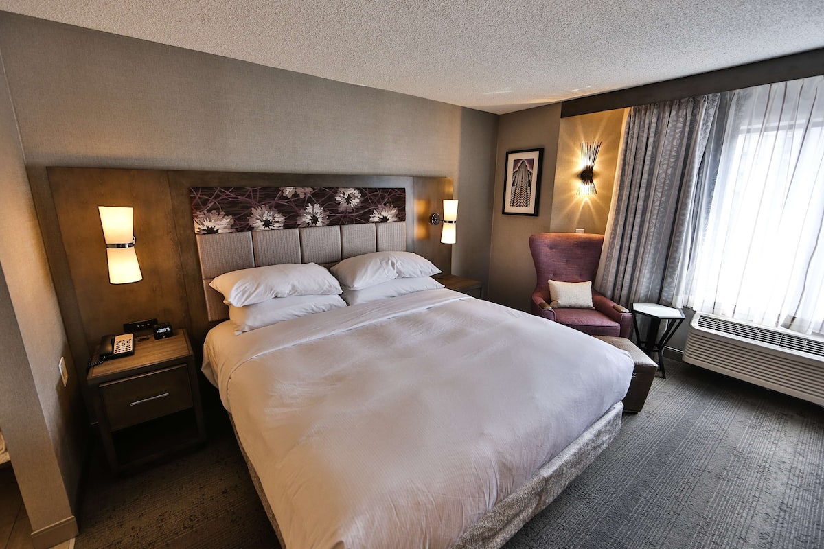 A hotel room with a large bed, a nightstand with a phone and lamp, a chair, a small table, and a window with curtains.