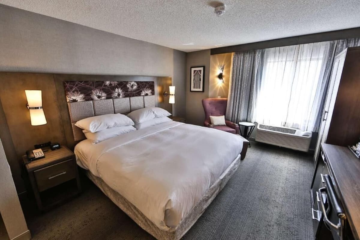 A hotel room with a large bed, bedside lamps, a chair, a desk, and a window with curtains.